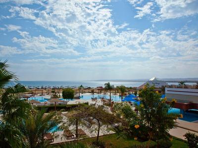 Blick auf Hotelpool mit Palmen, Strand und blauem Meer unter wolkigem Himmel.
