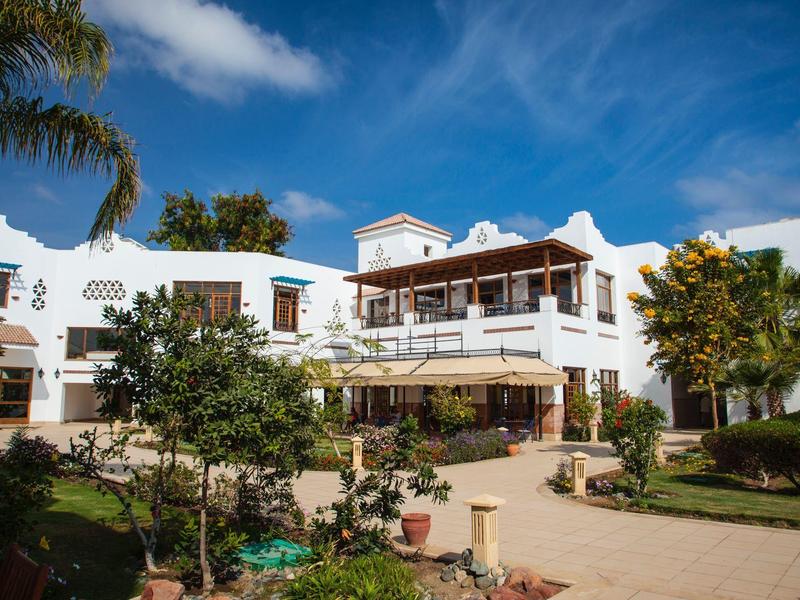 Bílá hotelová budova se zahradami a palmami pod jasně modrou oblohou.