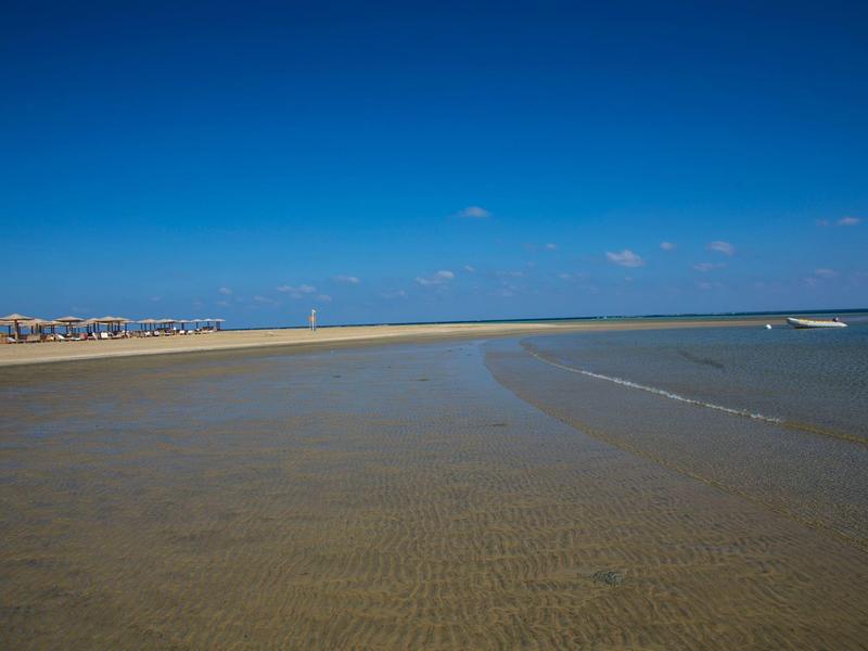 Rozlehlá pláž s jasným pískem a čistou modrou oblohou nad klidným mořem.