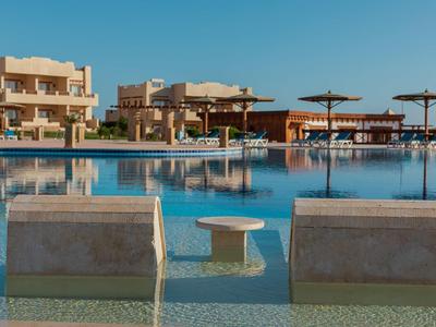 Großer Pool mit zwei halb eingetauchten Liegen und Sonnenschirmen vor Sandstein-Gebäuden.