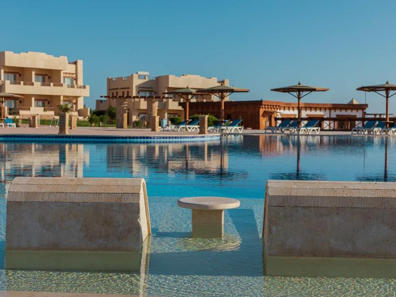 Großer Pool mit zwei halb eingetauchten Liegen und Sonnenschirmen vor Sandstein-Gebäuden.