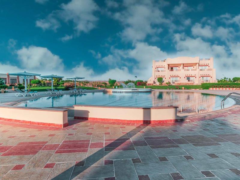 Großer runder Pool mit weißem Geländer vor einem hellen Gebäude unter blauem Himmel.