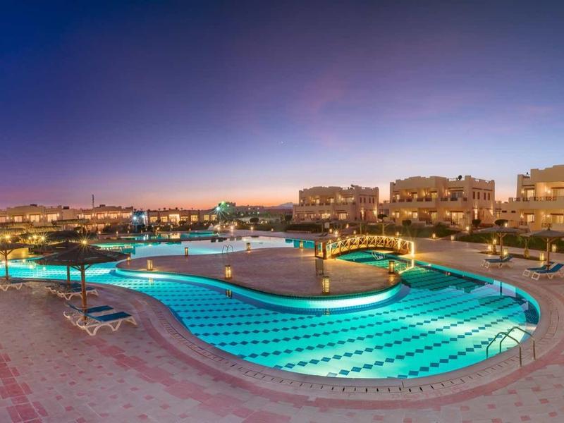 Nächtliches Panorama eines beleuchteten Hotels mit großem Pool und Sitzbereichen im Vordergrund.