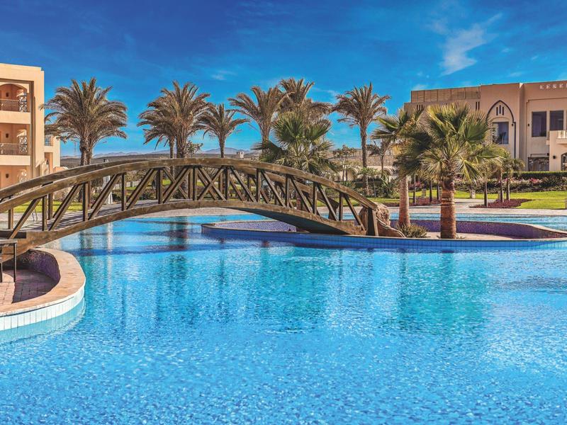 Large pool with bridge and palm trees in front of hotel buildings under blue sky.