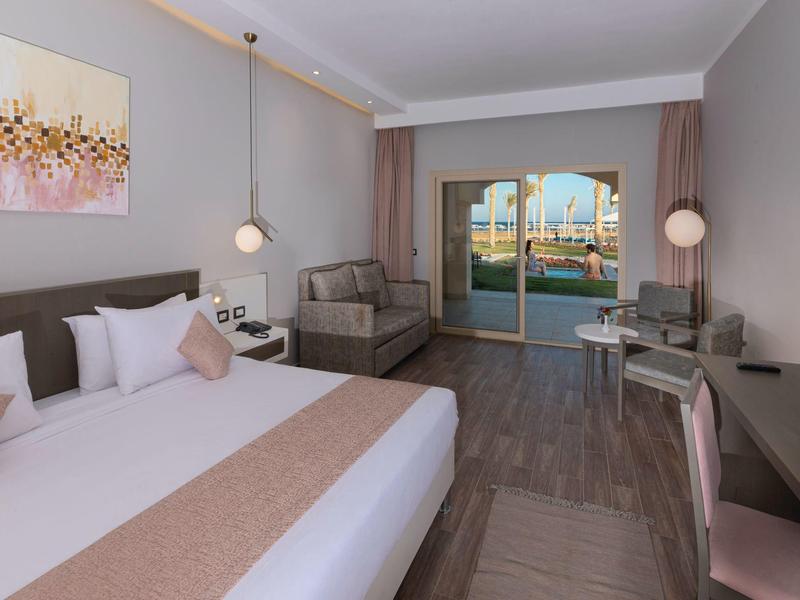 Modern hotel room with double bed, sofa, and balcony overlooking the water.