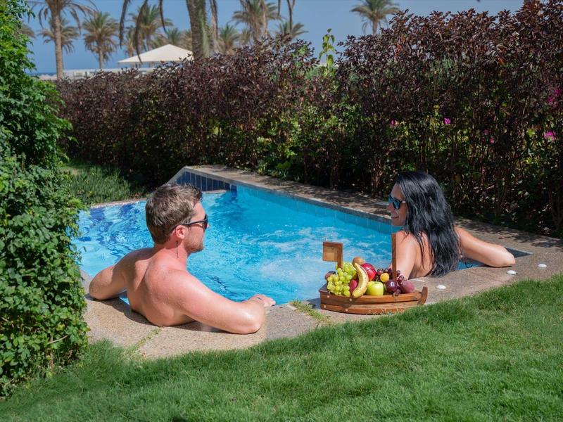 Two people relax in a pool next to a tray of fruit, surrounded by garden and hedges.