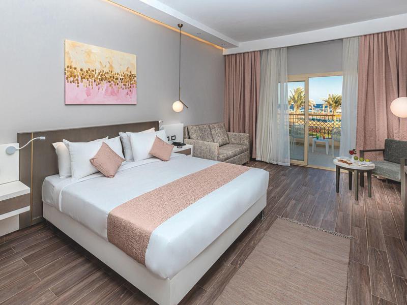 Modern hotel room with large bed, seating area, and balcony overlooking the water.