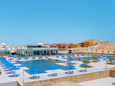 Großer Außenpool mit blauen Sonnenschirmen und Liegestühlen vor hellen Gebäuden unter klarem Himmel.