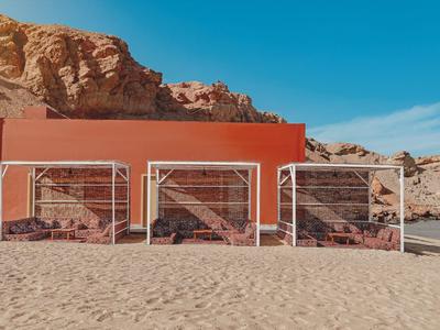 Drei kleine bunte Strandhütten mit Gittern vor felsigem Hintergrund und Sandboden.
