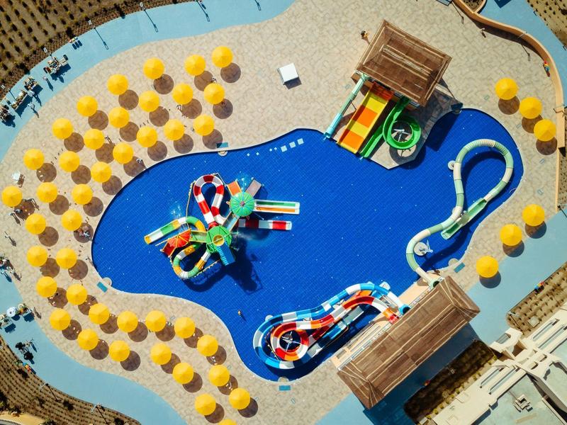 Freizeitbad mit blauem Becken, Wasserrutschen, zwei Holzterrassen und gelben Sonnenschirmen.