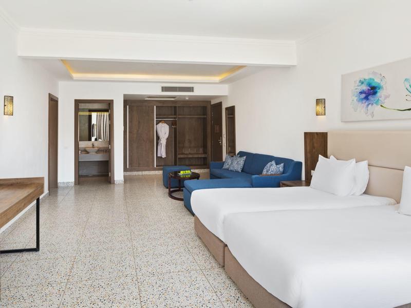 Helles Hotelzimmer mit zwei Doppelbetten, blauem Sofa, großem Schrank und Flachbildfernseher.