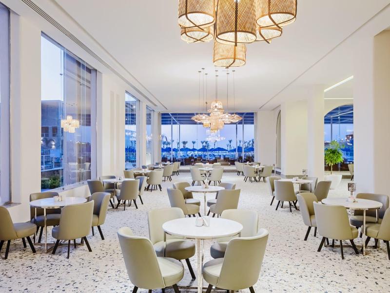 Modernes, helles Restaurant mit großen Fenstern, beigen Stühlen und runden weißen Tischen.