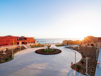 Przestronny teren hotelowy z okrągłą ścieżką, kwietnikiem na środku i widokiem na morze o zachodzie słońca.