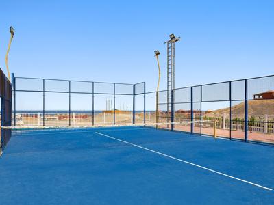 Zewnętrzny kort tenisowy z niebieską nawierzchnią i metalowym ogrodzeniem w słoneczny dzień.