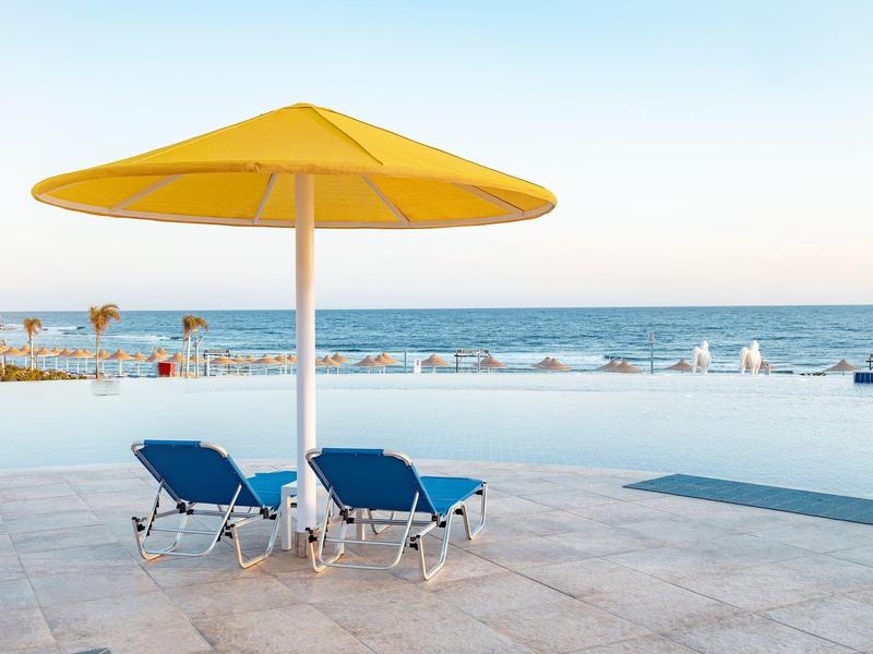 Dwa niebieskie leżaki pod żółtym parasolem na tle spokojnego morza.