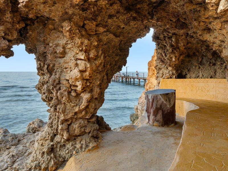 Widok na morze z naturalnego skalnego schronienia z wbudowaną ławką i pomostem w tle.