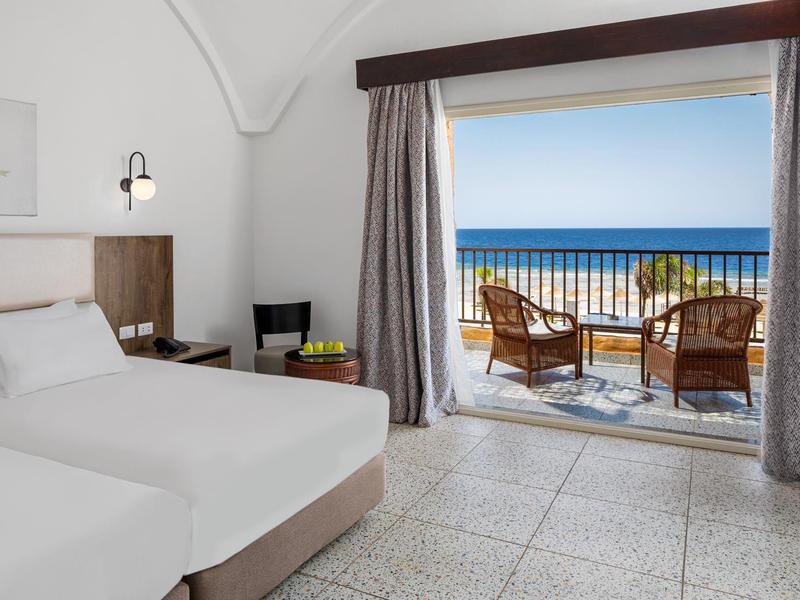 Elegancki pokój hotelowy z podwójnym łóżkiem, balkonem z widokiem na morze i drewnianymi krzesłami.