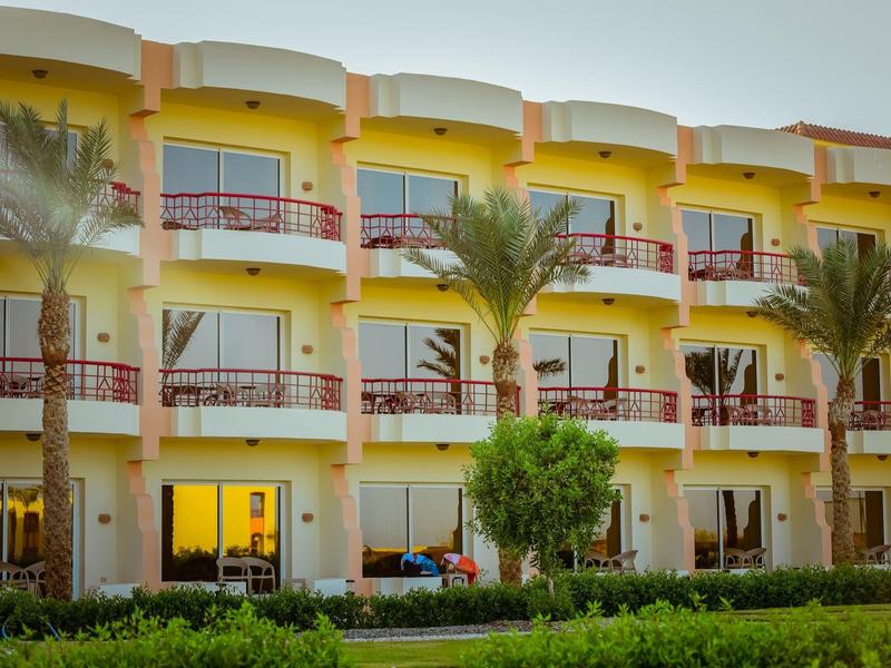 Mehrstöckiges gelb-weißes Hotel mit Balkonen, roten Geländern, Palmen und grünem Vorgarten.