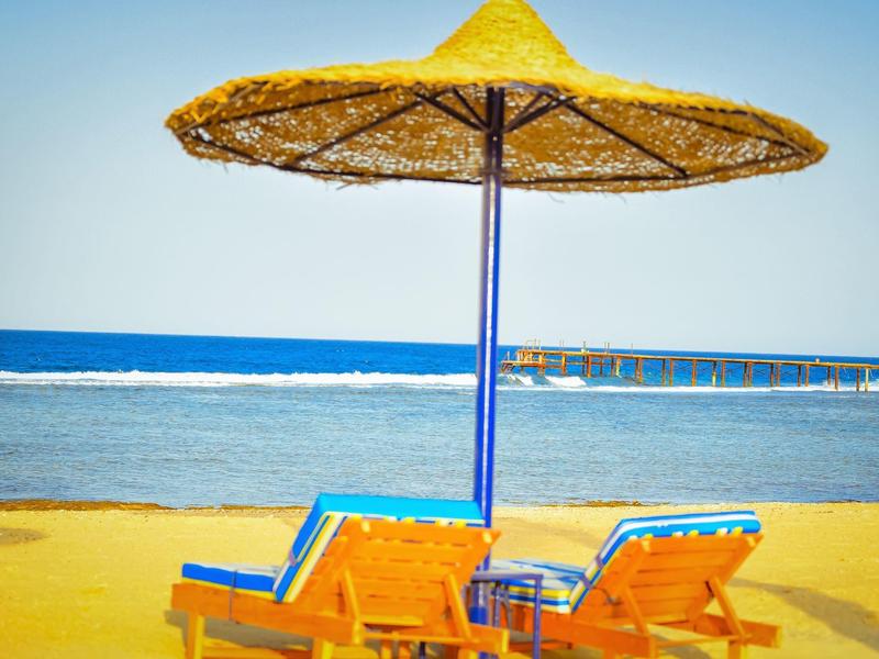 Zwei Holzliegen unter großem Strohsonnenschirm am Sandstrand mit Blick auf das blaue Meer.