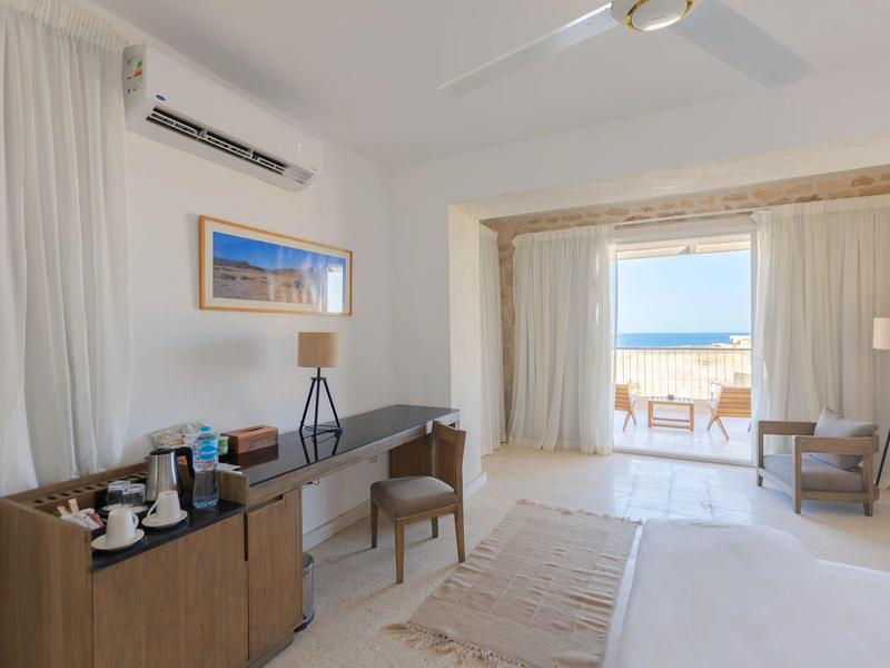 Habitación de hotel luminosa con zona de estar, escritorio y vista a la terraza con vistas al mar.