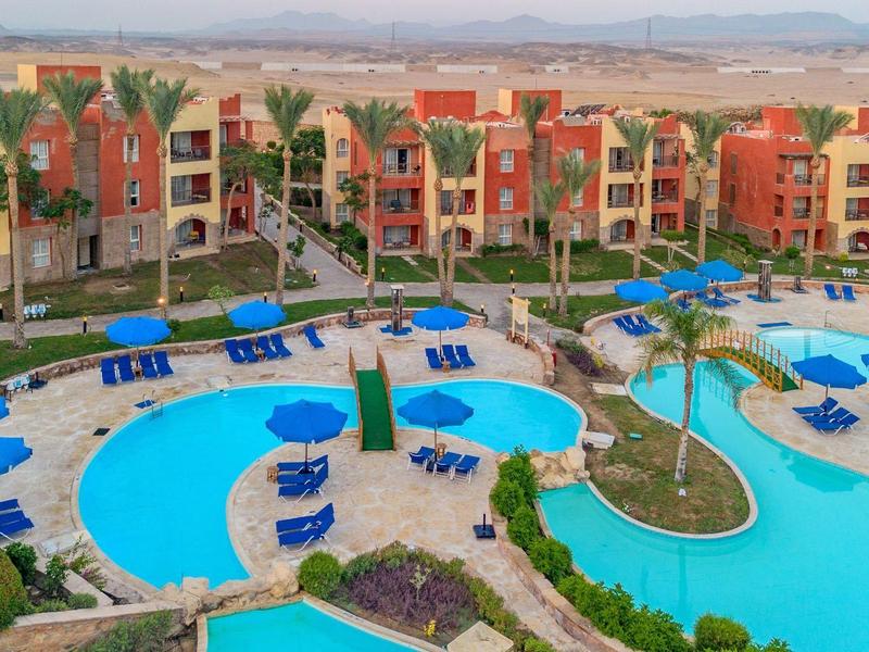 Gran piscina con tumbonas azules frente a edificios de hotel coloridos y montañas al fondo.
