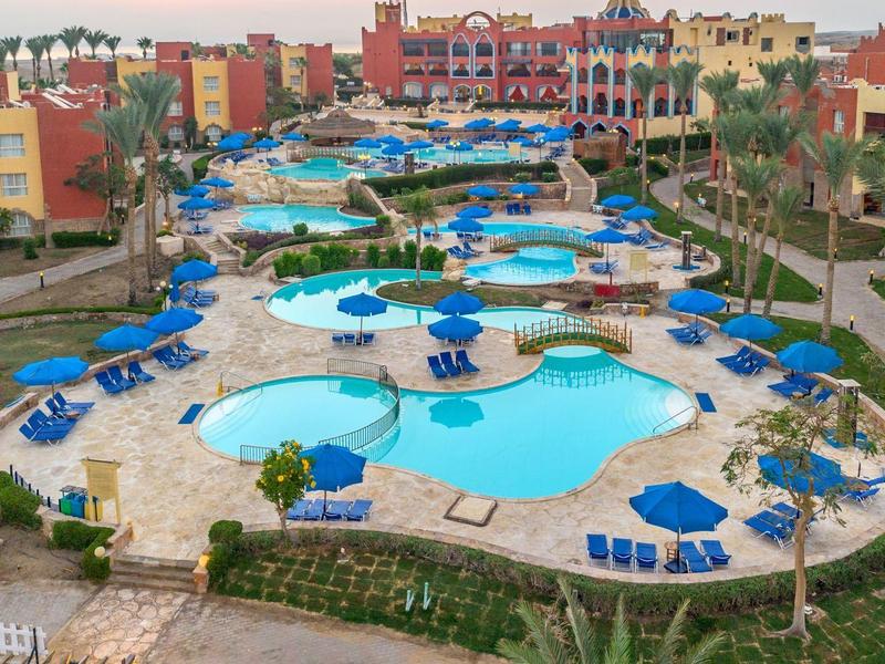 Gran área de piscinas con varias piscinas, sombrillas azules y edificios coloridos al fondo