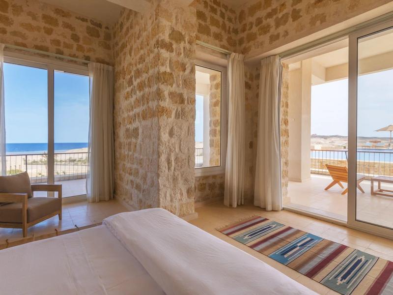 Habitación de hotel con paredes de piedra, vista al mar y terraza con sillas y sombrilla.