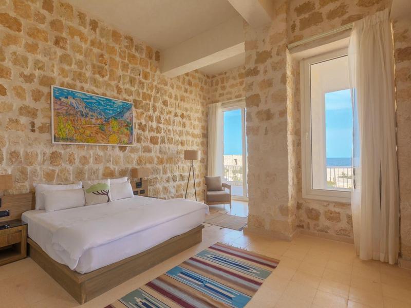 Dormitorio luminoso con paredes de piedra, cama grande y vista al mar a través de puertas abiertas.