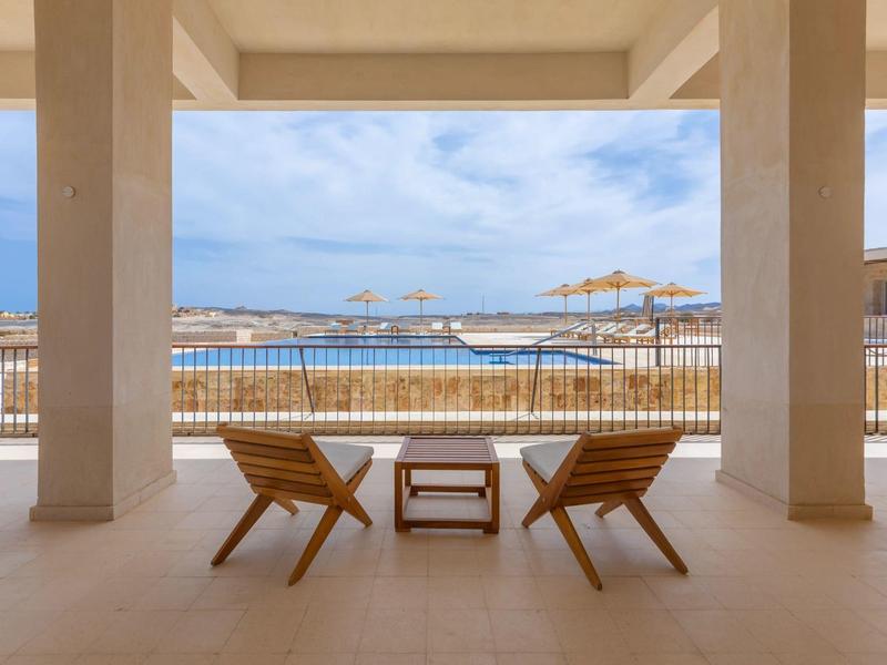 Dos tumbonas con una mesa pequeña en una terraza con vista al mar y una playa con sombrillas.