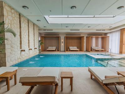 Ein heller Innenpoolraum mit blauer Wasserfläche, Liegen und Holzstühlen, dekoriert mit Pflanzen.