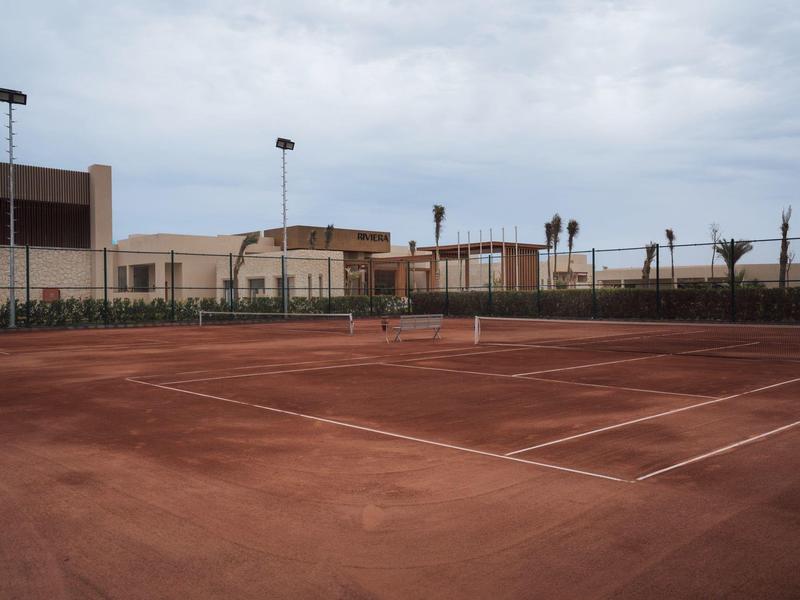 Court de tennis en terre battue vide avec des bâtiments en arrière-plan sous un ciel nuageux.