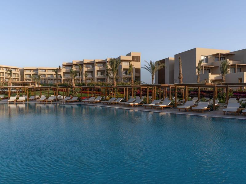 Grand piscine d'hôtel avec chaises longues et bâtiment hôtelier moderne en arrière-plan sous un ciel clair.