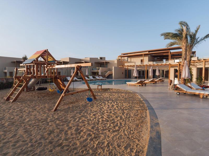 Aire de jeux avec balançoires et structure d'escalade à côté d'une piscine et de bâtiments d'hôtel au coucher du soleil.