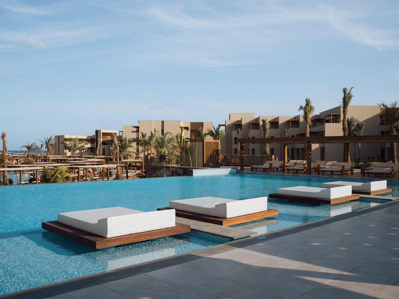 Piscine extérieure moderne avec lits blancs et plusieurs bâtiments en arrière-plan sous un ciel clair.