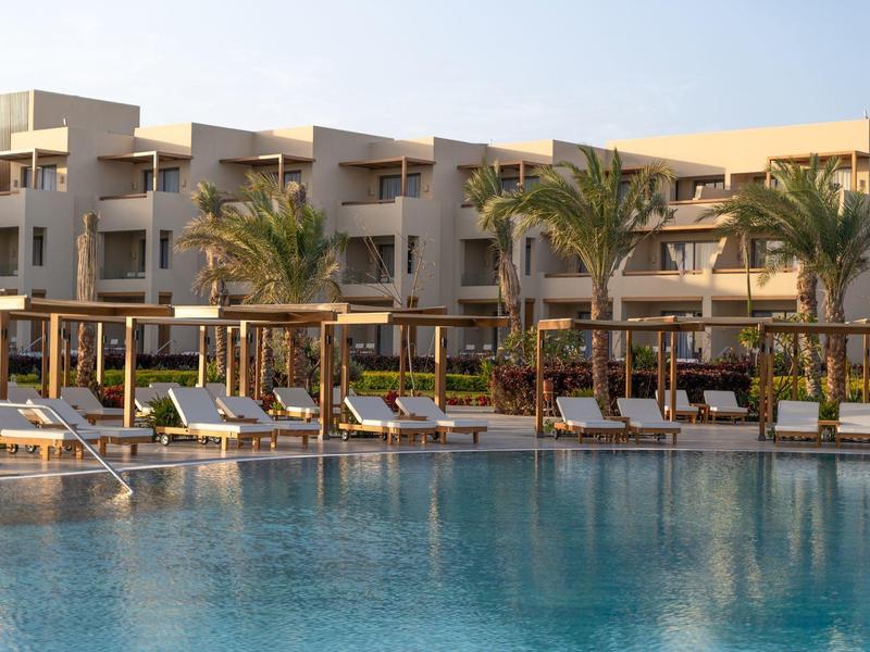 Complexe hôtelier moderne avec piscine, transats et palmiers sous un ciel clair.