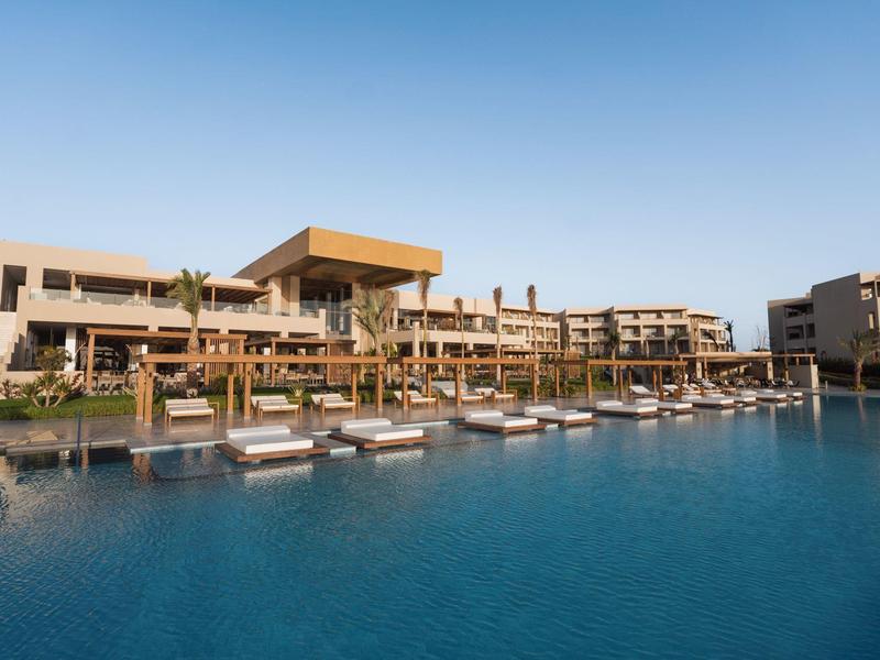 Hôtel moderne avec grande piscine, transats et ciel clair en arrière-plan.