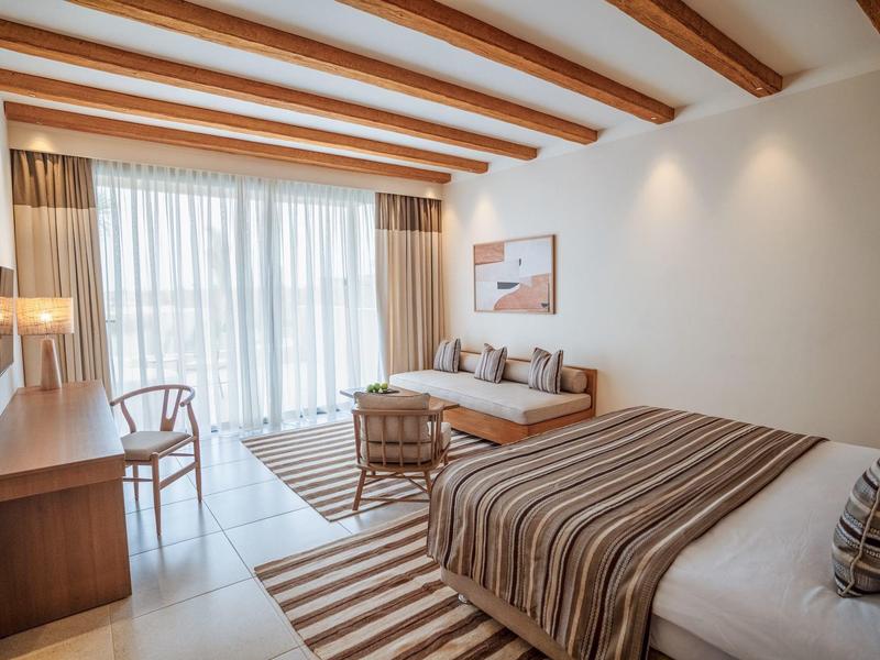 Chambre d'hôtel lumineuse avec lit double, canapé, bureau et grande fenêtre avec rideaux.