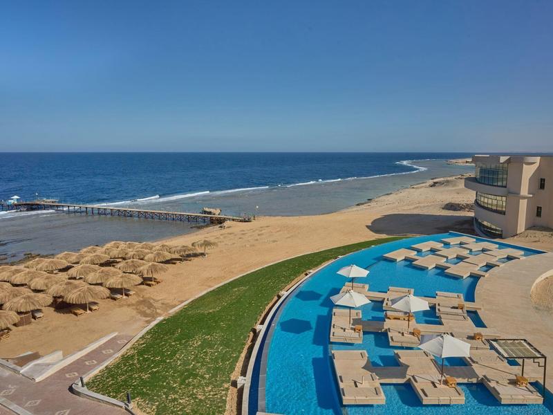 View of the beach with umbrellas and lounge chairs next to a pool at a hotel resort.