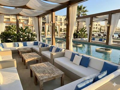 Salon confortable avec meubles en bois et coussins à côté d'une piscine sous une pergola.