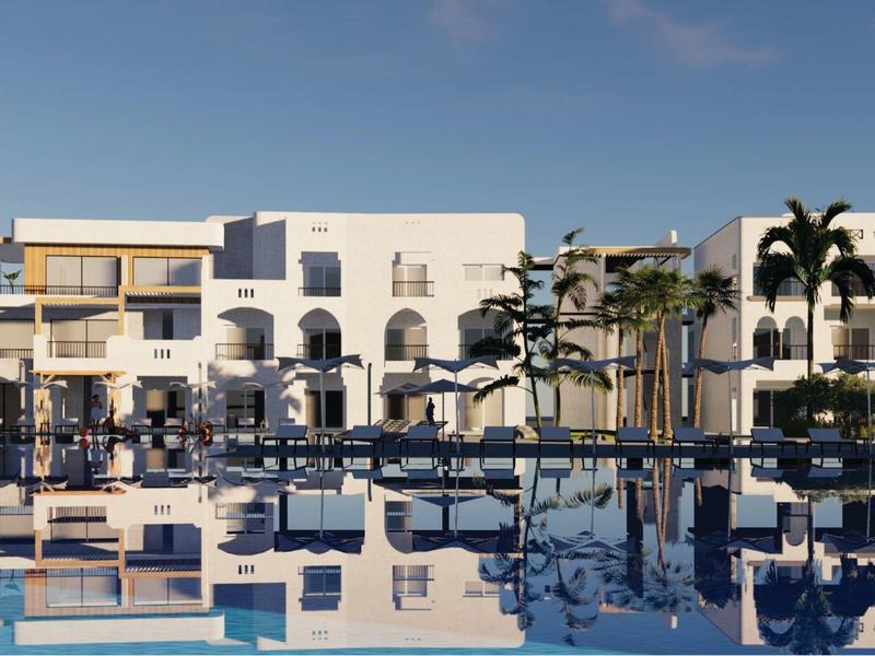 A modern multi-story hotel with palm trees and a calm pool in the foreground under a clear sky.