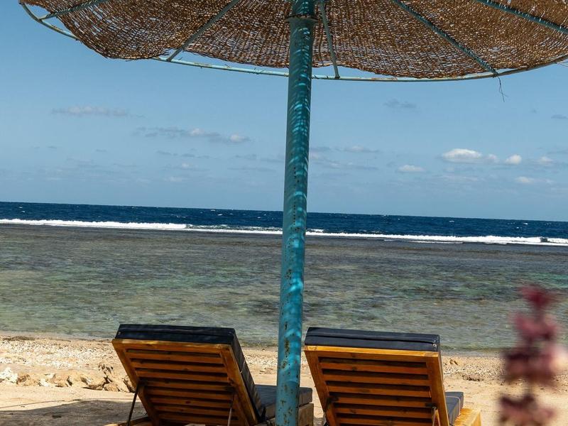 Deux chaises longues en bois sous un grand parasol en paille sur une plage de sable avec vue sur la mer.
