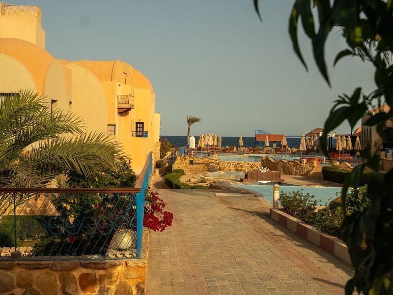 Chemin d'hôtel ensoleillé avec bâtiments jaunes, palmiers et piscine en arrière-plan.