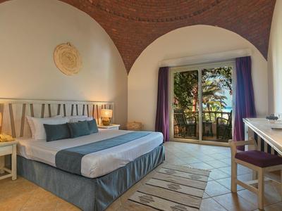 Jasny pokój hotelowy z dużym łóżkiem, lampkami nocnymi i balkonem z roślinami.