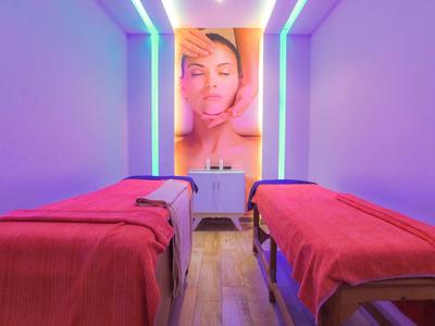 Dwa łóżka do zabiegów z czerwonymi kocami w pokoju z fioletowym oświetleniem i dużym plakatem na ścianie.