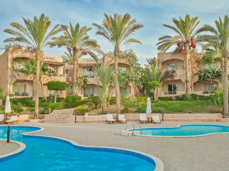 Budynek hotelu z wieloma pokojami, basenem i palmami na tle słonecznego dnia.