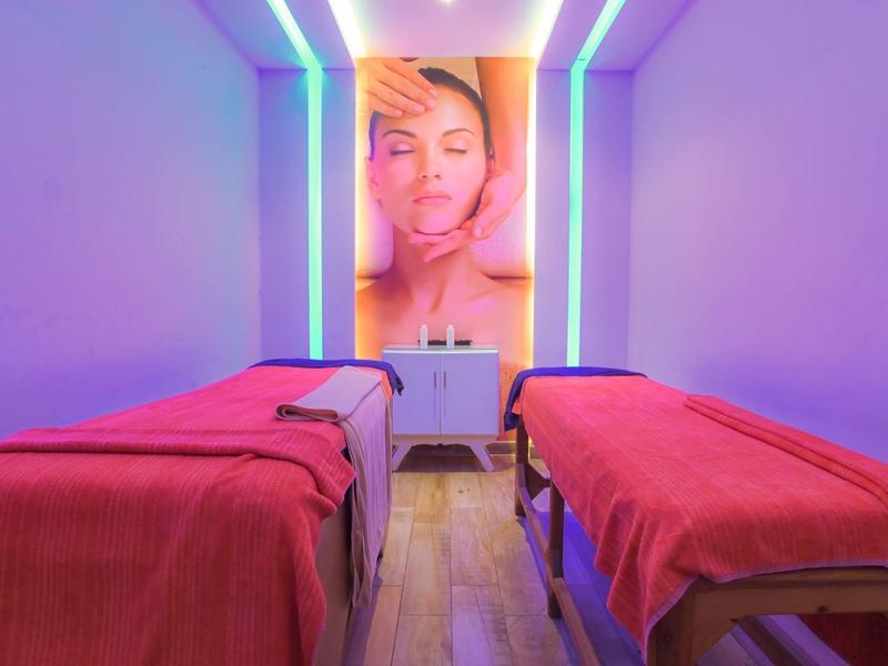 Dwa łóżka do zabiegów z czerwonymi kocami w pokoju z fioletowym oświetleniem i dużym plakatem na ścianie.