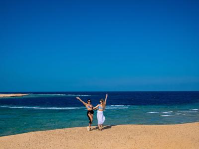 Zwei Personen tanzen freudig am Sandstrand mit blauem Himmel und Meer im Hintergrund.