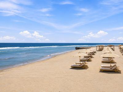 Strand mit blauem Himmel, ruhigem Meer und Reihen von Liegestühlen unter Sonnenschirmen.