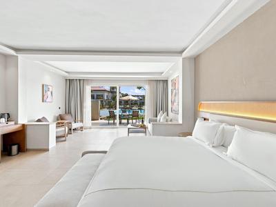 Hell eingerichtetes Hotelzimmer mit großem Bett und Blick auf einen Balkon mit Meerblick.