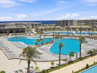 Großer Hotelpool mit Liegestühlen und Palmen unter blauem Himmel am Meer.